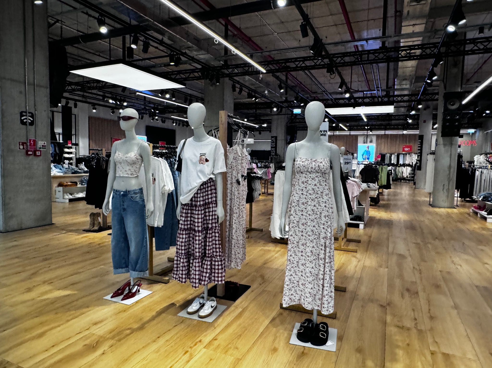 Wnętrze sklepu odzieżowego: trzy manekiny prezentują stylizacje z letniej kolekcji na tle drewnianej podłogi i regałów z ubraniami. Widoczne oświetlenie sufitowe.