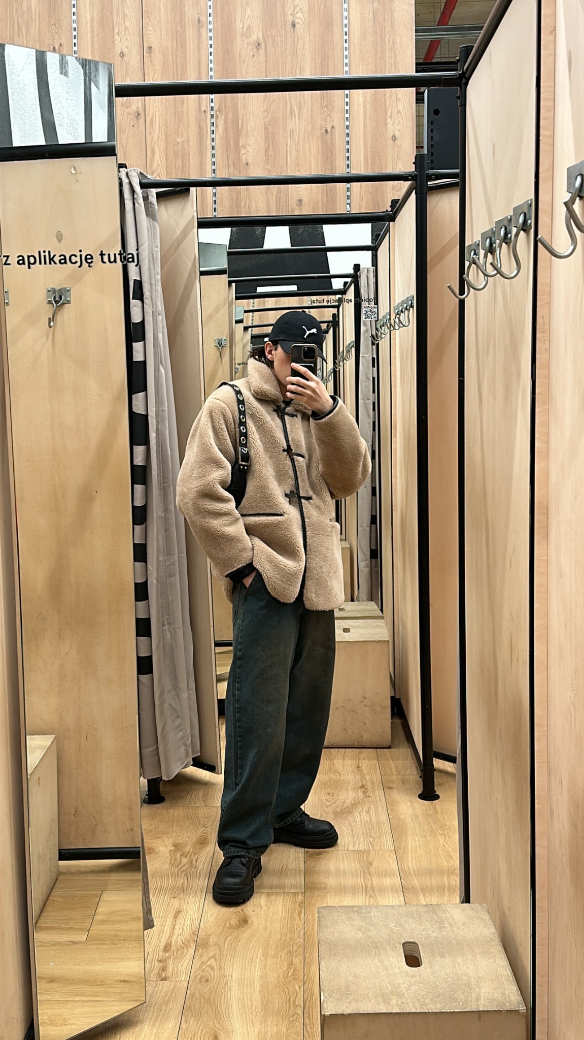 Osoba w beżowej kurtce typu baranek, czapce z daszkiem i jeansach robi selfie w przymierzalni z drewnianymi ścianami i metalowymi wieszakami. Widoczne lustro i podest.