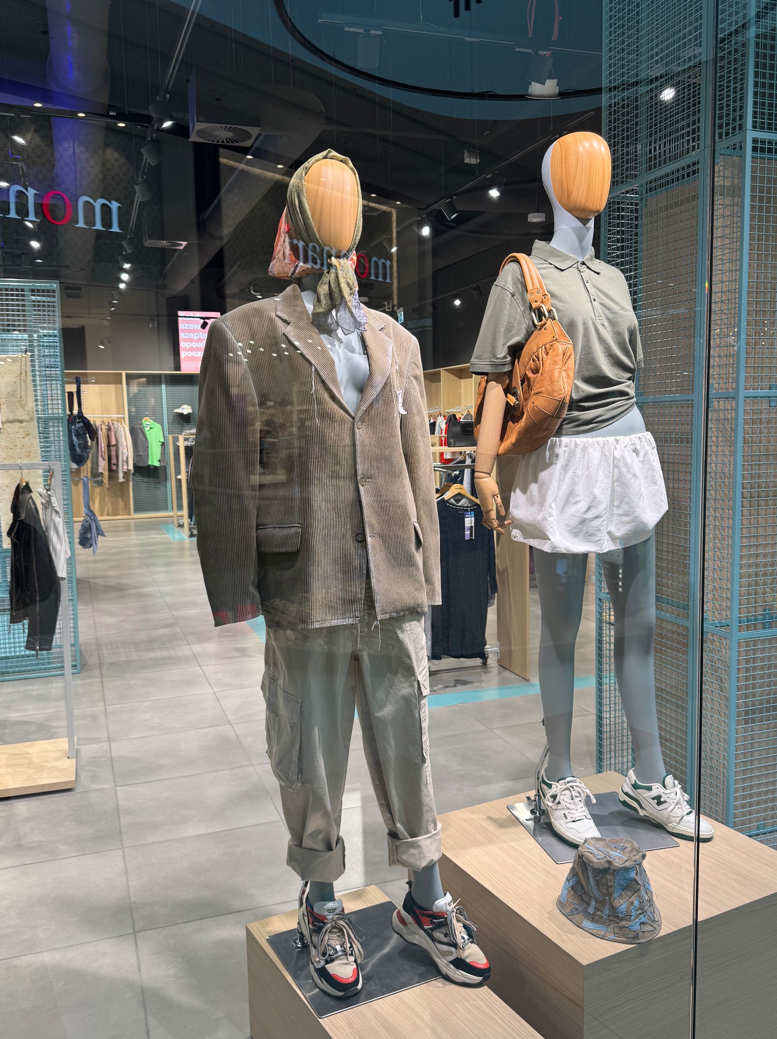 Dwa manekiny w witrynie sklepowej prezentują stylizacje: jeden w brązowym sztruksowym garniturze i sportowych butach, drugi w polo, spódniczce i torbie na ramię.