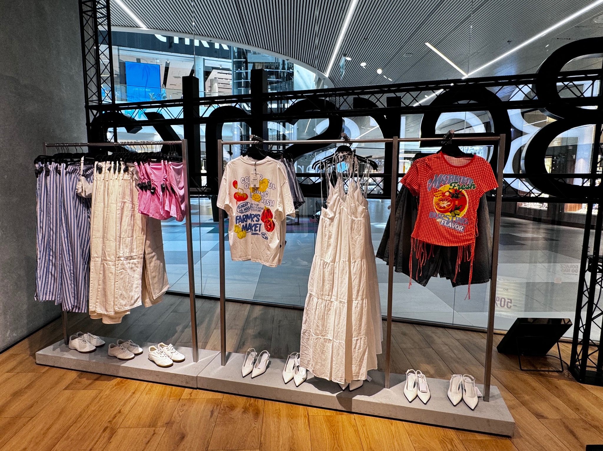 Ekspozycja odzieży w sklepie: sukienka maxi, koszulki z nadrukami, spodnie i szorty na wieszakach, dopełnione białymi butami i szpilkami. Nowoczesne wnętrze sklepu.