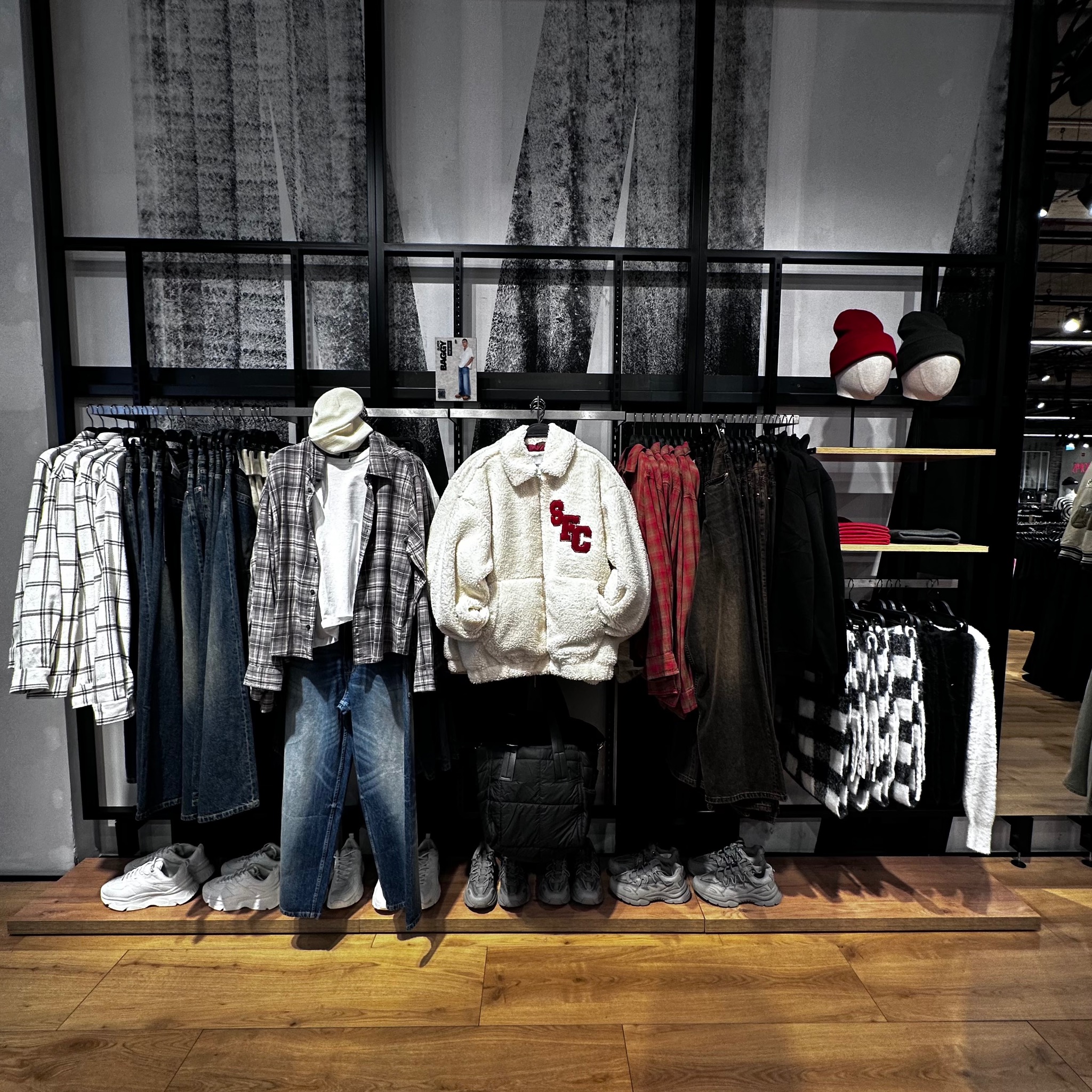 Ekspozycja odzieży w sklepie: flanelowe koszule, jeansy, kurtka z futerka, czapki na manekinach. Ułożone na półce buty sportowe. Minimalistyczny design.