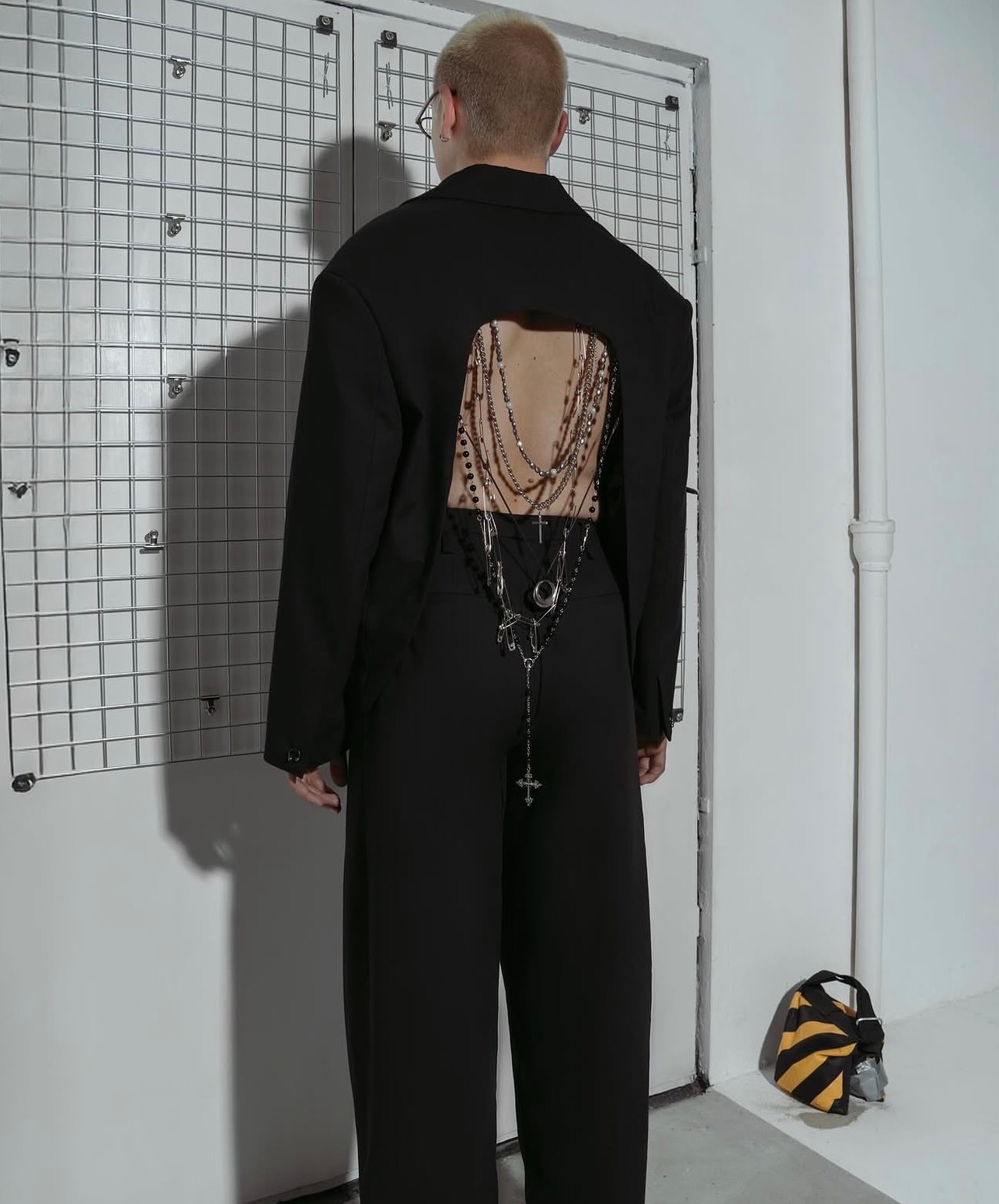 Osoba w czarnym, awangardowym garniturze z wycięciem na plecach, ozdobionym łańcuchami i krzyżami. Widok z tyłu, minimalistyczne tło, torba w paski leży na podłodze.