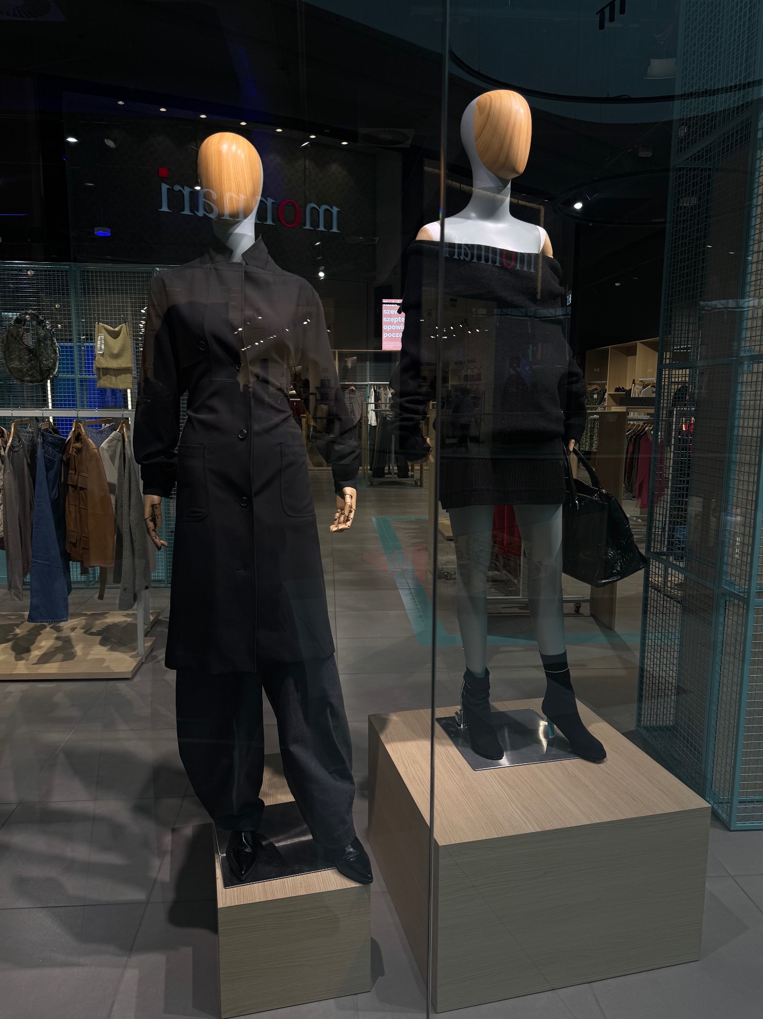 Dwa manekiny w czarnych stylizacjach w witrynie sklepowej. Jeden w płaszczu, drugi w sukience z odkrytymi ramionami i torebką. Minimalistyczny design i nowoczesny wystrój.