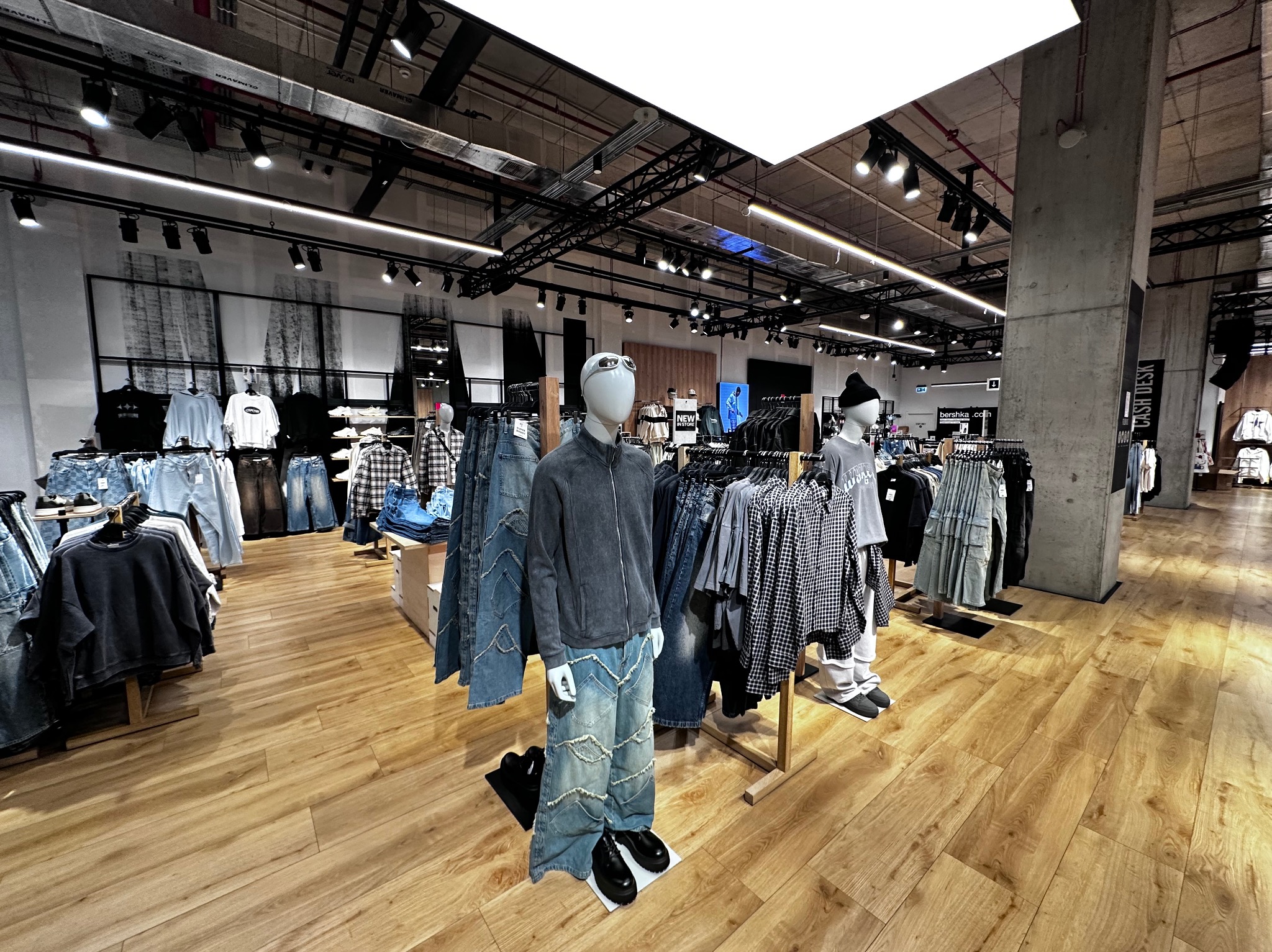 Jasne wnętrze sklepu odzieżowego z manekinami prezentującymi najnowsze kolekcje. Widoczne ubrania na wieszakach i drewniana podłoga. Oświetlenie sufitowe.