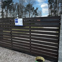 Ogrody U Karola - Ciemnobrązowe, poziome deski ogrodzenia posesji z tabliczką informacyjną firmy ochroniarskiej. Obok stoi wiadro z farbą. Tło: drzewa i błękitne niebo.