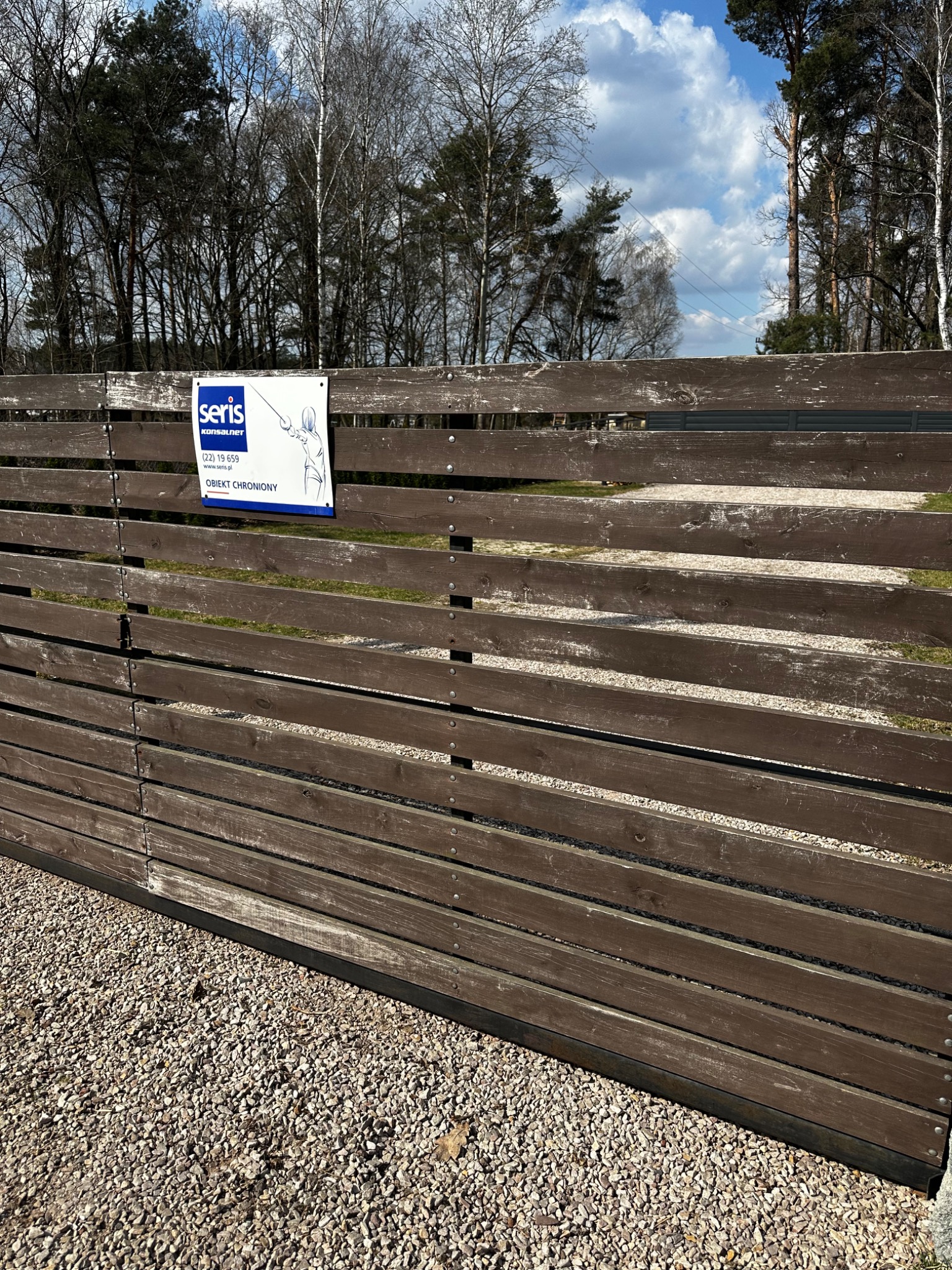 Drewniany płot posesji z tabliczką informującą o ochronie obiektu, widok na zewnątrz, w tle drzewa i błękitne niebo z chmurami.