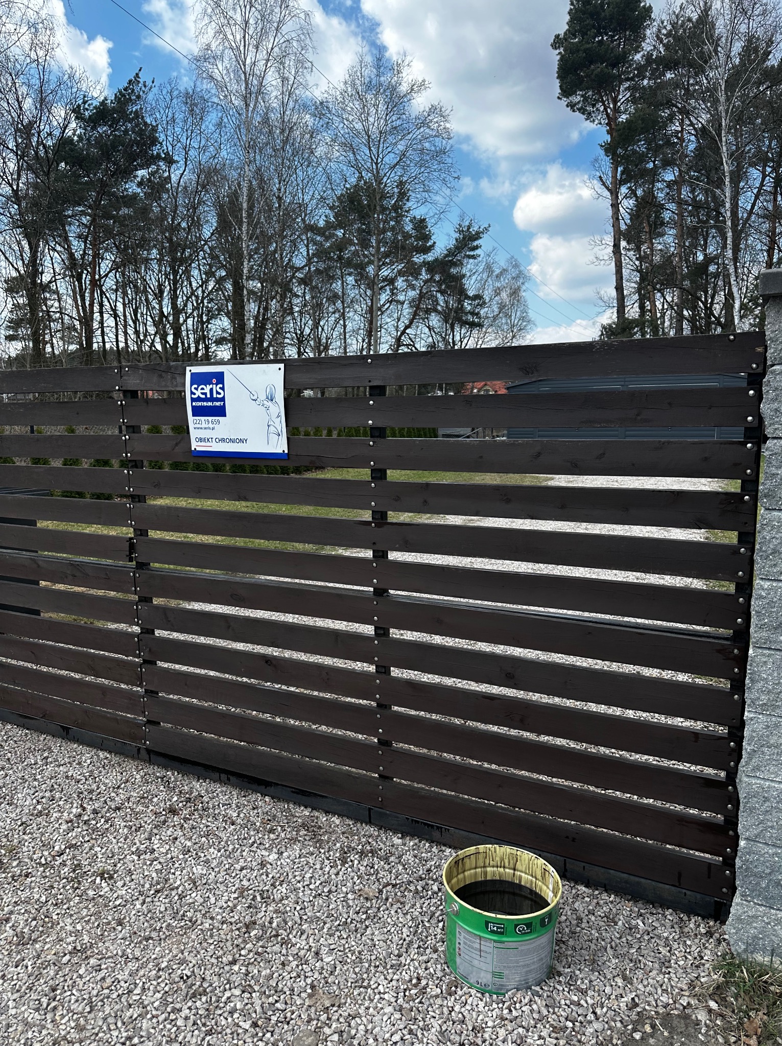 Ciemnobrązowe, poziome deski ogrodzenia posesji z tabliczką informacyjną firmy ochroniarskiej. Obok stoi wiadro z farbą. Tło: drzewa i błękitne niebo.