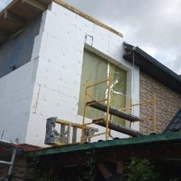SolidHouse - Ocieplanie ściany budynku styropianem, widoczne rusztowanie i fragment dachu. Prace w toku, widok z dołu na elewację w trakcie izolacji.