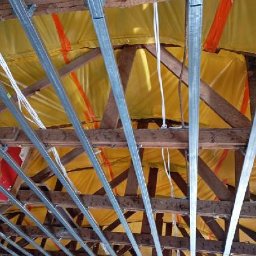 SolidHouse - Sufit z drewnianą konstrukcją i metalowymi profilami, zabezpieczony żółtą folią. Widoczne białe kable. Ujęcie z dołu, perspektywa zbieżna.