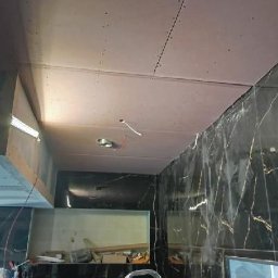 SolidHouse - Montaż płyt karton-gips na suficie w pomieszczeniu z marmurowymi ścianami. Widoczna drabina i narzędzia. Prace wykończeniowe w trakcie realizacji.