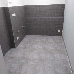 SolidHouse - Wykończona łazienka z szarymi płytkami na ścianach i podłodze. Widoczne gniazdka elektryczne i bateria. Minimalistyczny design.