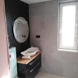 SolidHouse - Wykończona łazienka w Sulęcinie z umywalką na szafce z drewnianym blatem, szarą glazurą i czarną ścianą z geometrycznym wzorem. Lustro owinięte folią ochronną.