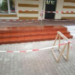 SolidHouse - Świeżo pomalowane, ceglane schody wejściowe do budynku, zabezpieczone taśmą ostrzegawczą. Widoczny drewniany kozioł. Jasna elewacja z poziomymi pasami.