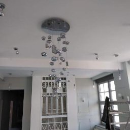 SolidHouse - Jasne wnętrze po remoncie z nowoczesnym oświetleniem: chromowany żyrandol kulisty i punktowe lampy sufitowe. Widoczne okna i fragment rusztowania.