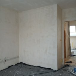 Luk bud - Pokój w trakcie szpachlowania: ściany pokryte gładzią, podłoga zabezpieczona folią ochronną. Widoczne narzędzia i materiały budowlane w tle.