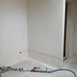 Luk bud - Wnętrze po szpachlowaniu: gładkie, jasne ściany w rogu pokoju, widoczny otwór drzwiowy i szlifierka do gipsu z odkurzaczem na podłodze. Przygotowanie do malowania.