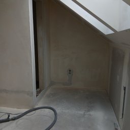Luk bud - Wnętrze poddasza po szpachlowaniu, widoczne ściany przygotowane do malowania, fragment okna dachowego i rura odkurzacza przemysłowego na podłodze.