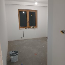 Luk bud - Pokój po szpachlowaniu i malowaniu na biało, widoczne narzędzia i puszka farby. Okno w drewnianej ramie. Gładkie ściany, przygotowane do dalszych prac wykończeniowych.