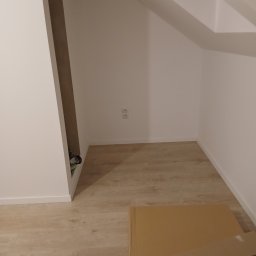 Luk bud - Poddasze ze skosem, wykończone w bieli, z jasną podłogą. Widoczne gniazdka elektryczne, fragment zabudowy i kartony na podłodze. Minimalistyczne wykończenie.