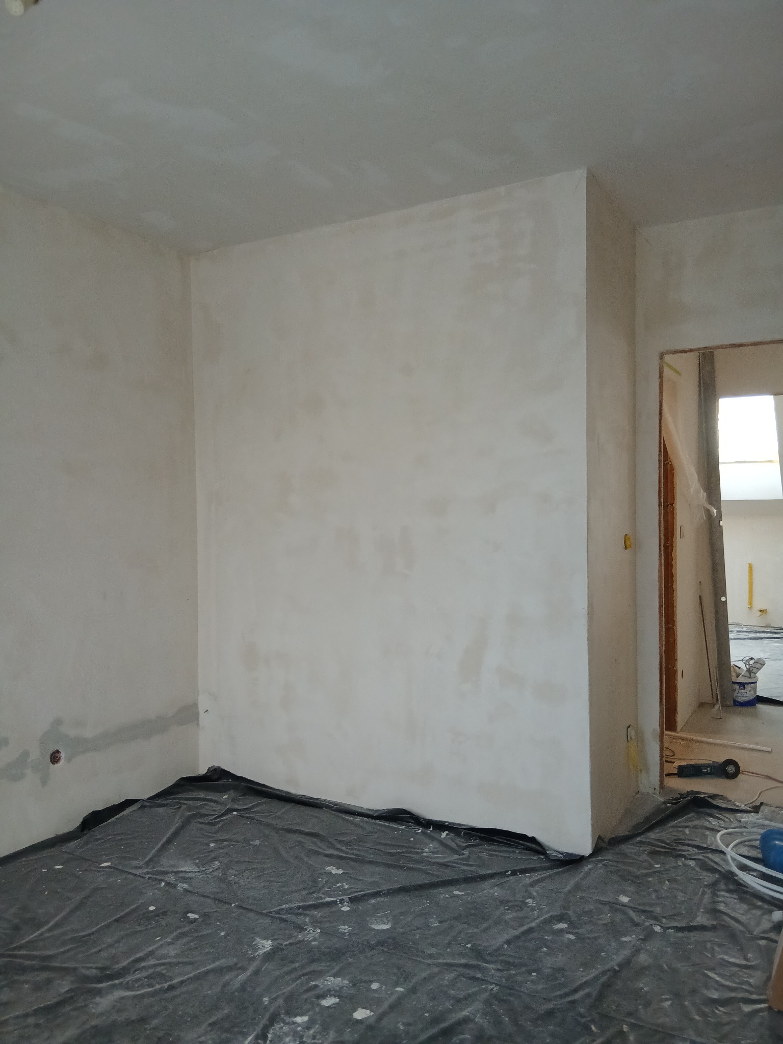 Pokój w trakcie szpachlowania: ściany pokryte gładzią, podłoga zabezpieczona folią ochronną. Widoczne narzędzia i materiały budowlane w tle.