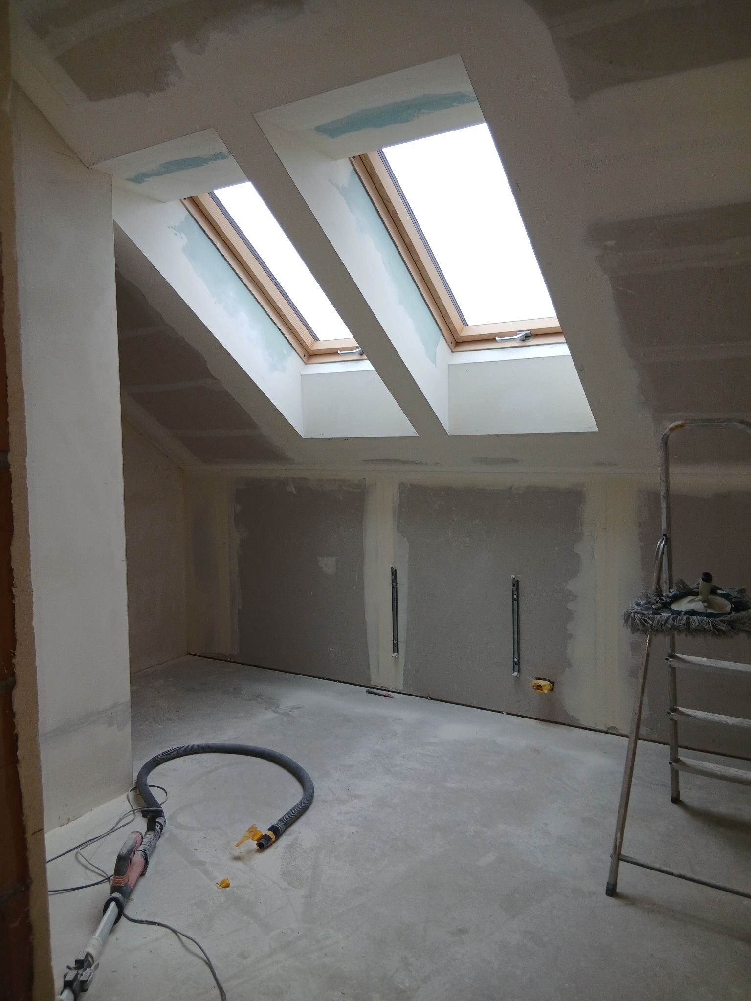 Poddasze w trakcie szpachlowania z oknami dachowymi, widoczne narzędzia i drabina. Ściany przygotowane pod malowanie, surowy beton na podłodze.
