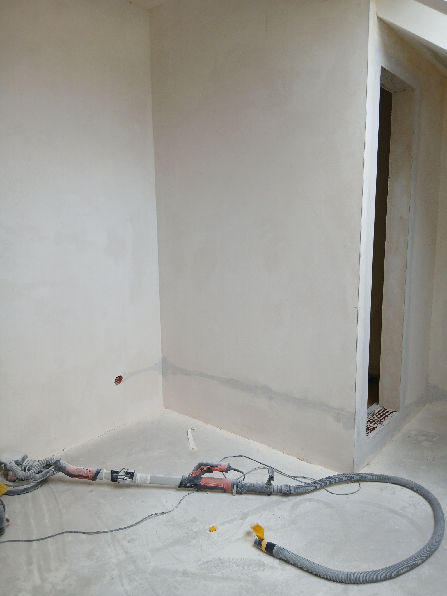 Wnętrze po szpachlowaniu: gładkie, jasne ściany w rogu pokoju, widoczny otwór drzwiowy i szlifierka do gipsu z odkurzaczem na podłodze. Przygotowanie do malowania.