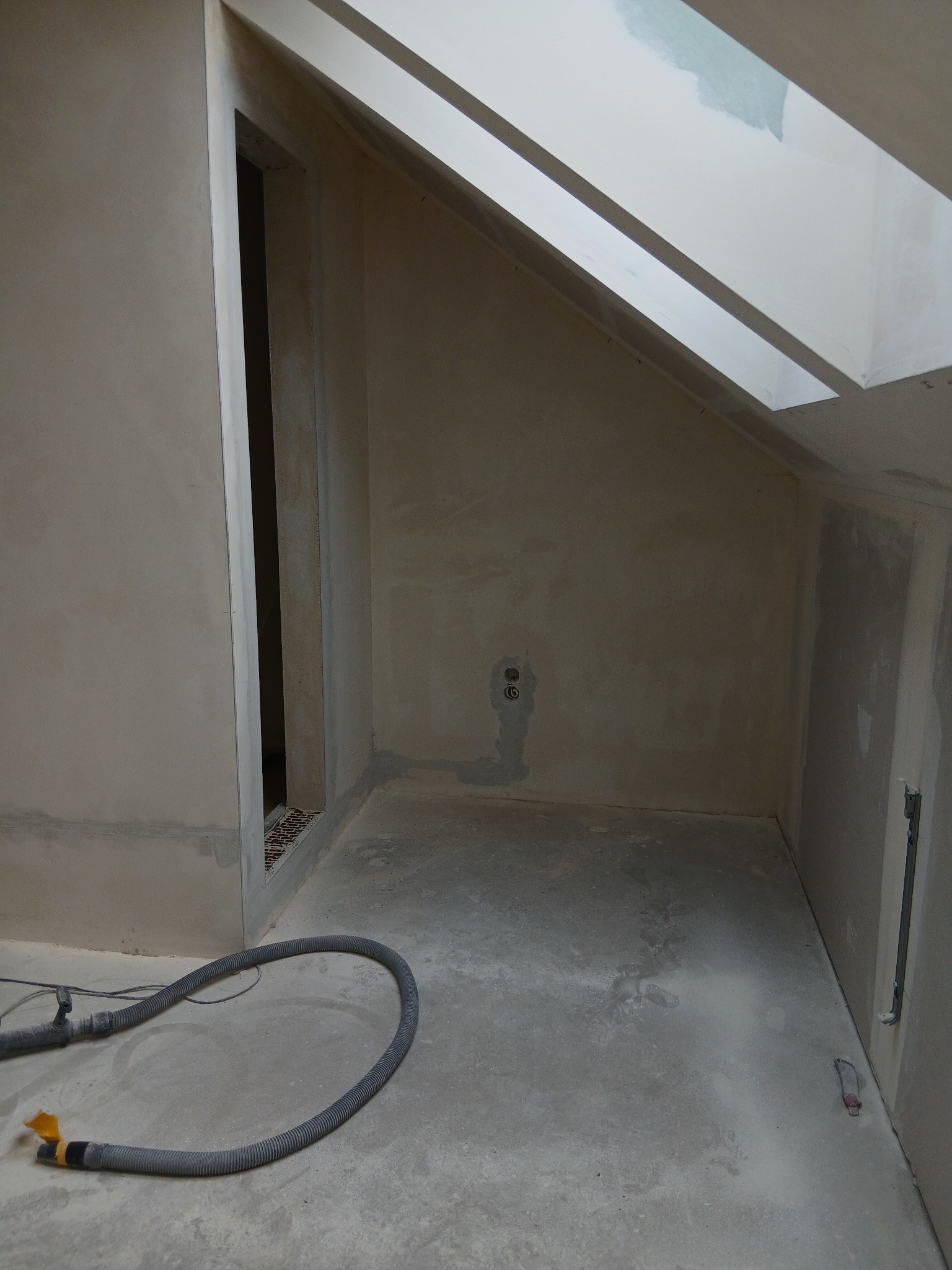 Wnętrze poddasza po szpachlowaniu, widoczne ściany przygotowane do malowania, fragment okna dachowego i rura odkurzacza przemysłowego na podłodze.