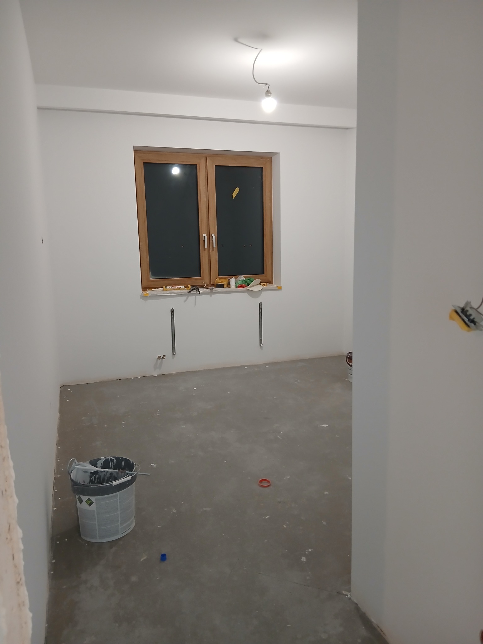 Pokój po szpachlowaniu i malowaniu na biało, widoczne narzędzia i puszka farby. Okno w drewnianej ramie. Gładkie ściany, przygotowane do dalszych prac wykończeniowych.