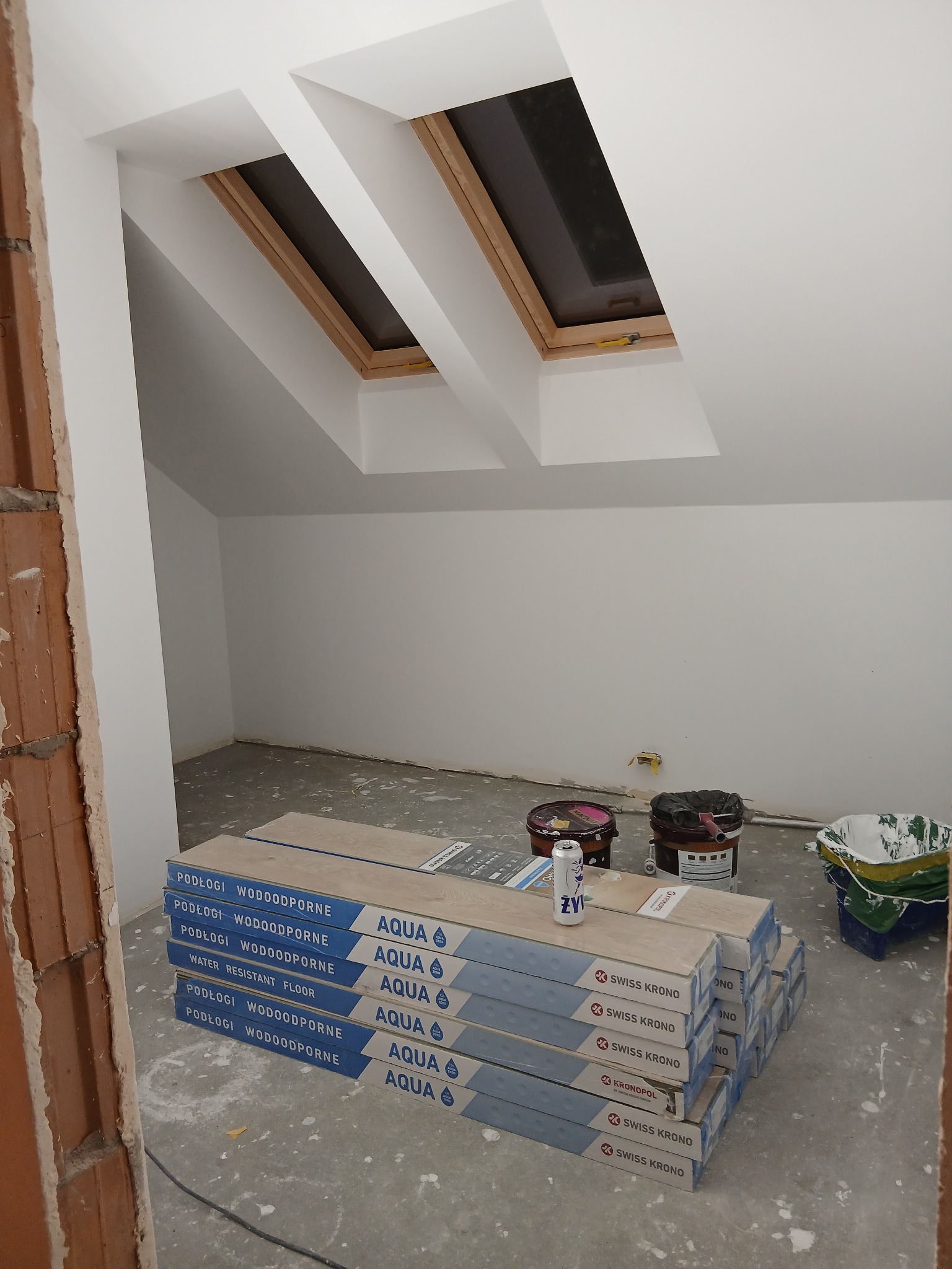 Poddasze w trakcie wykańczania: ułożone paczki paneli podłogowych Swiss Krono Aqua, widoczne okna dachowe i ściany przygotowane do malowania.
