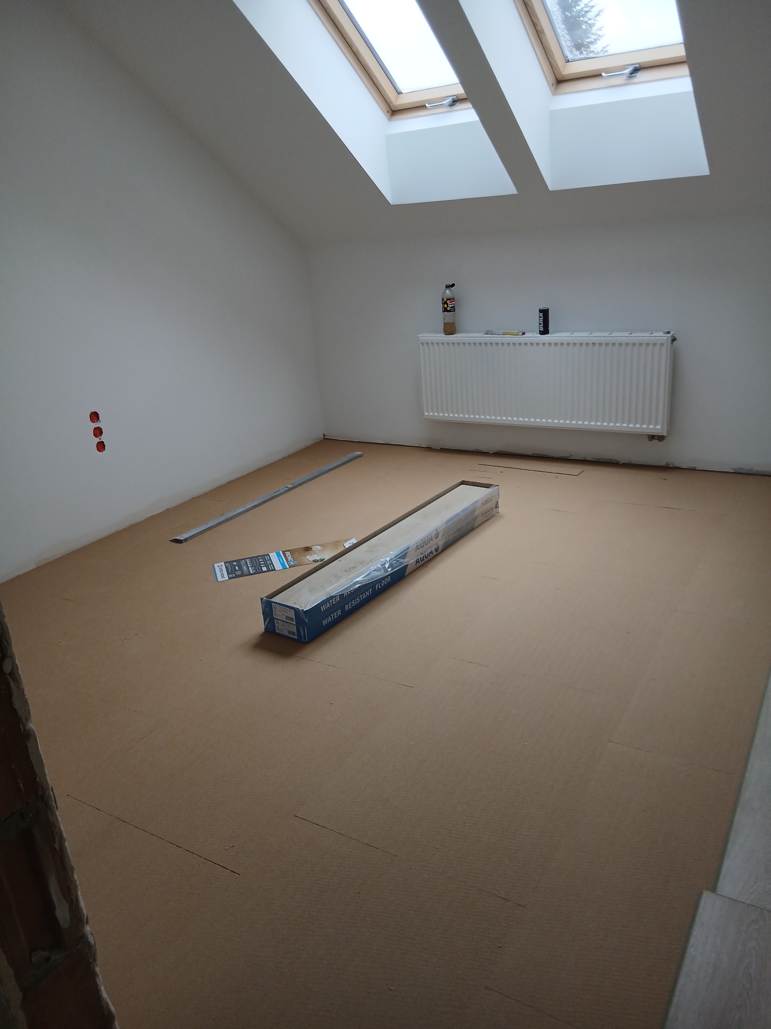 Podkład pod panele ułożony w pokoju na poddaszu z oknami dachowymi. Widoczna paczka paneli, listwa przypodłogowa i grzejnik. Przygotowanie do montażu podłogi.