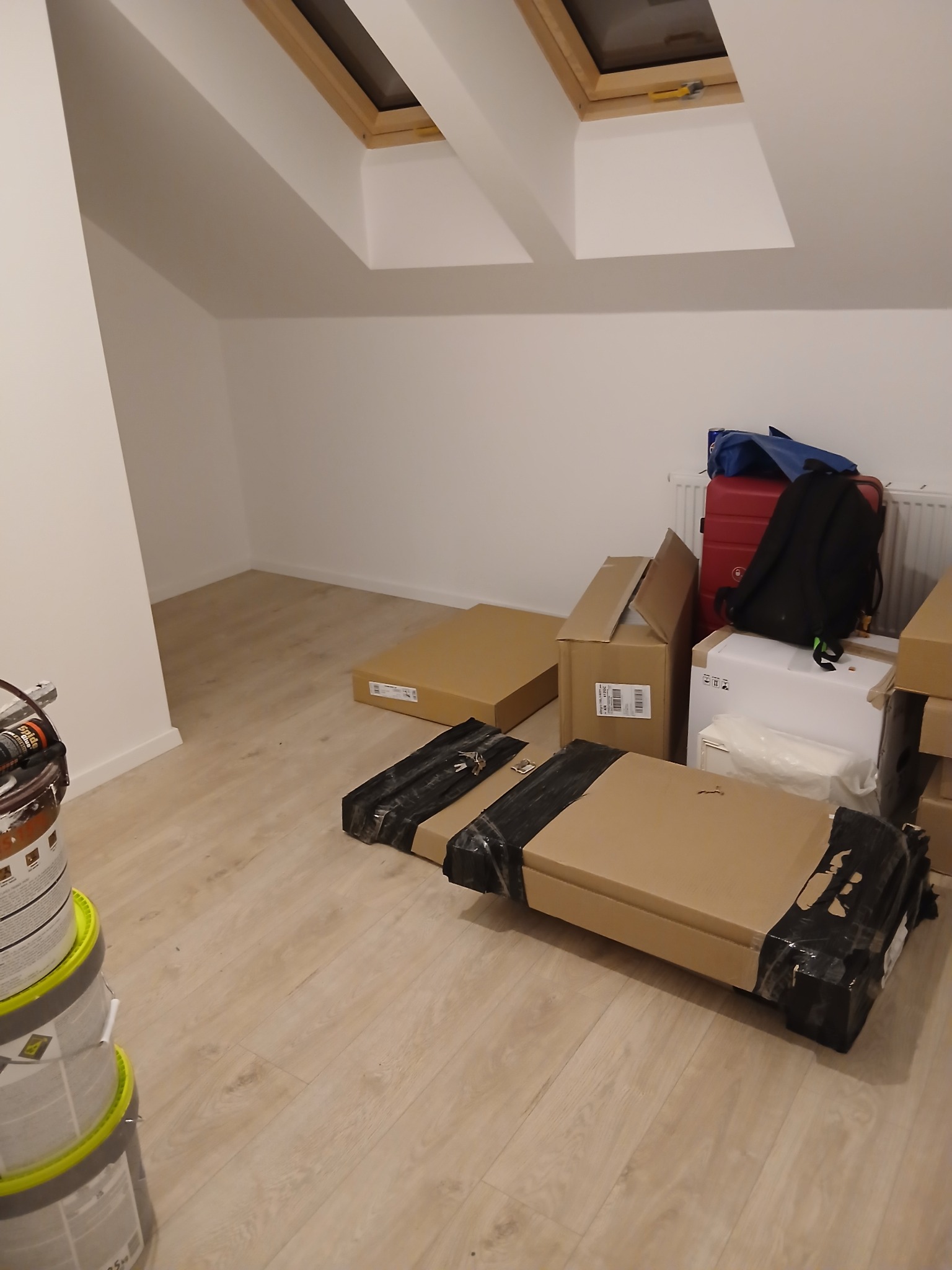 Poddasze z oknami dachowymi, przygotowane do prac wykończeniowych. Widoczne farby, kartony z meblami i bagaże. Podłoga wyłożona panelami.
