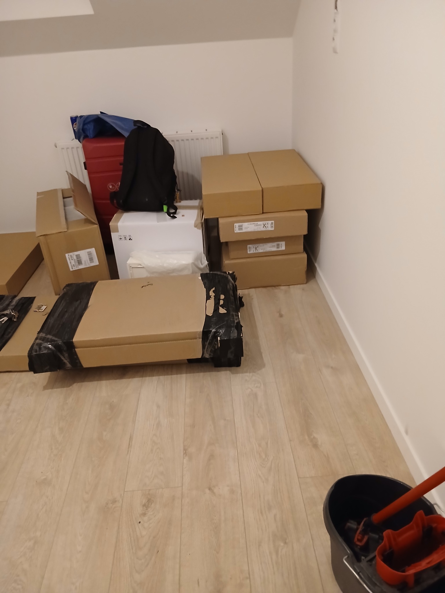 Kartonowe paczki i torba w rogu pokoju z panelami podłogowymi. Przy ścianie wiadro z mopem. Przygotowania do remontu lub przeprowadzki.