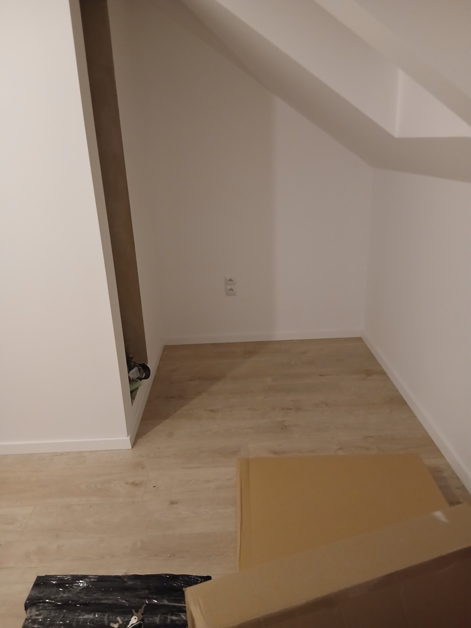 Poddasze ze skosem, wykończone w bieli, z jasną podłogą. Widoczne gniazdka elektryczne, fragment zabudowy i kartony na podłodze. Minimalistyczne wykończenie.