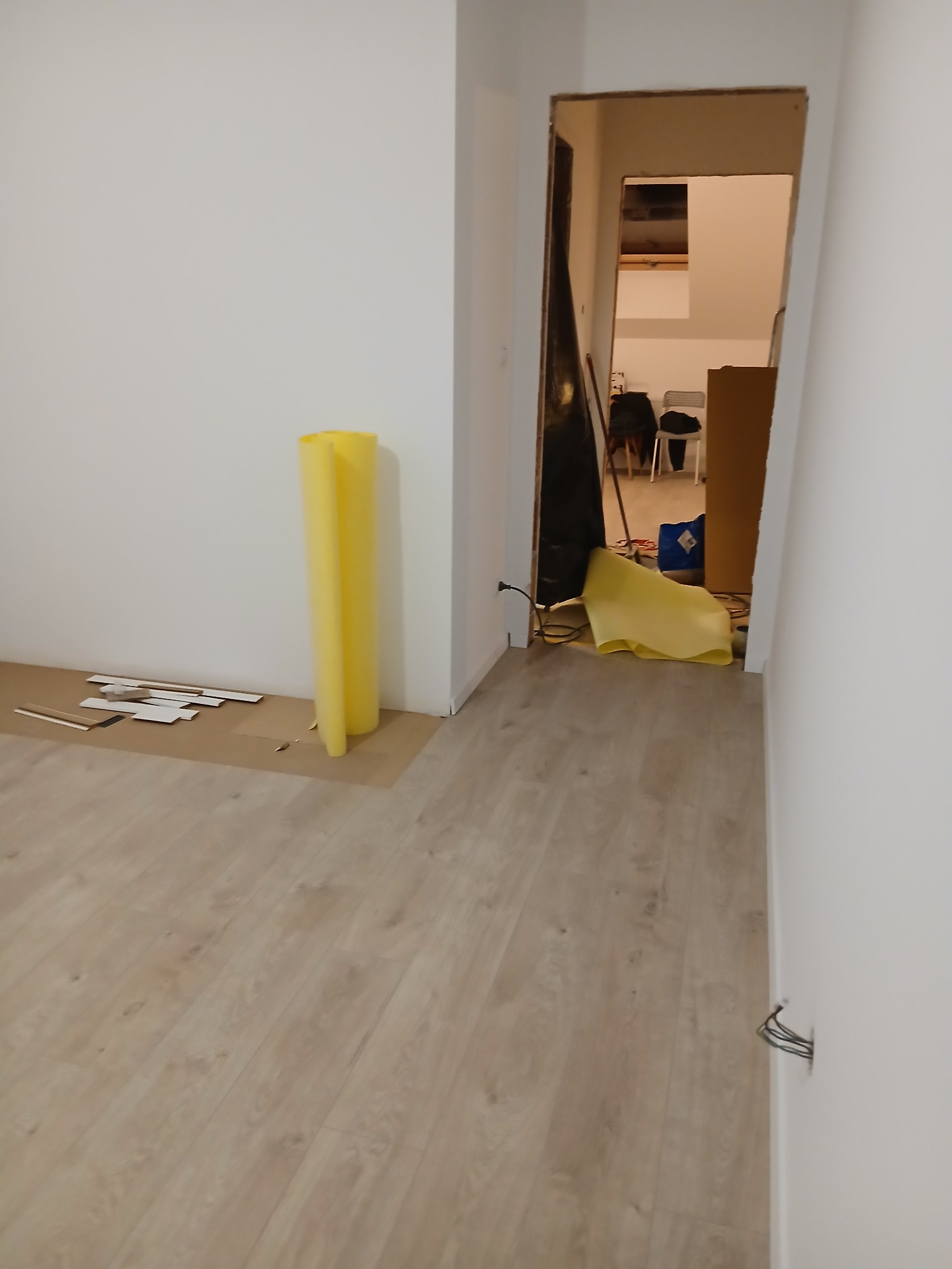 Jasne panele podłogowe w trakcie montażu, widoczne listwy przypodłogowe i żółta folia ochronna. W tle otwarte przejście do innego pomieszczenia.