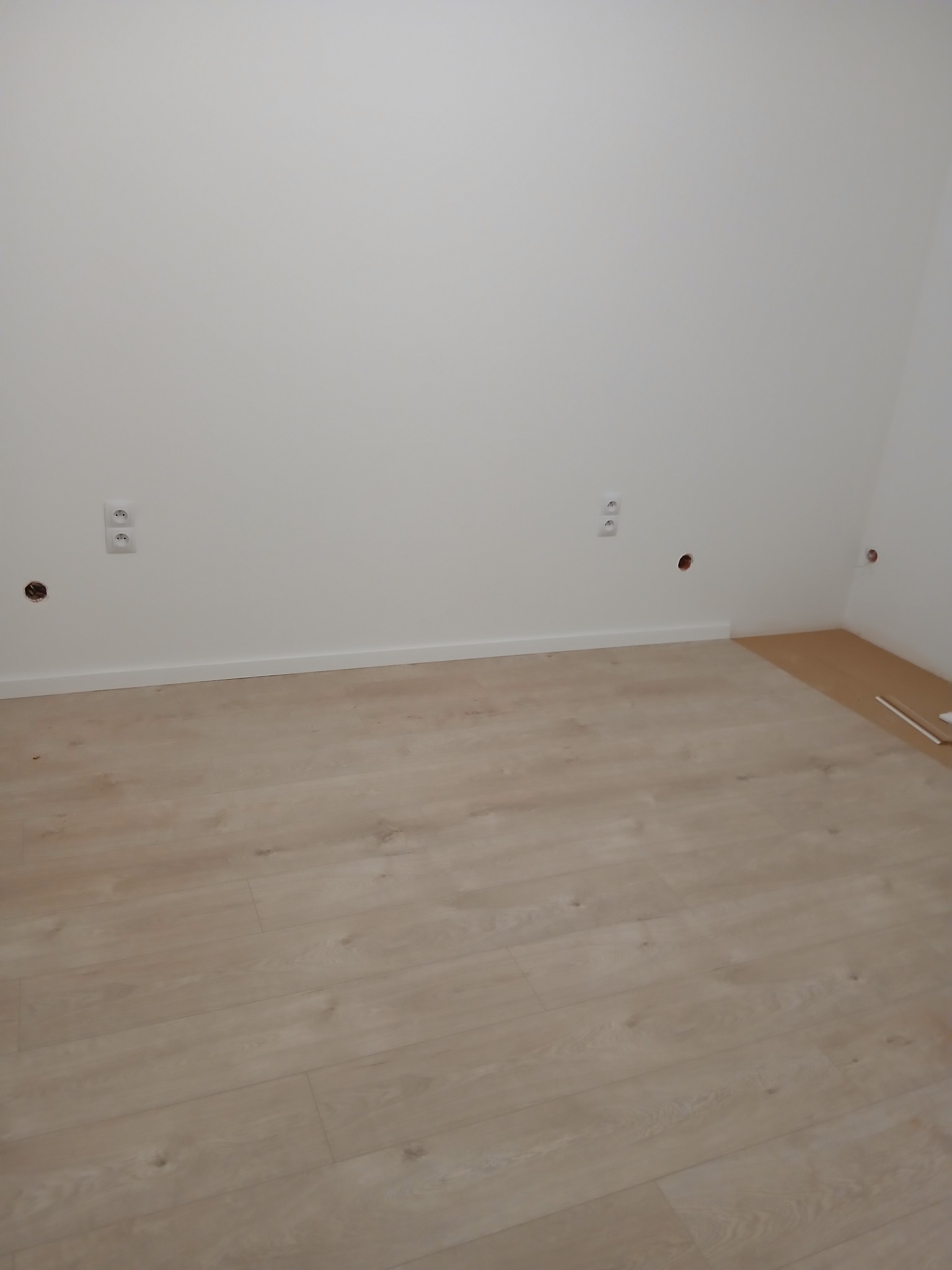 Jasne panele podłogowe w nowym pokoju z białymi ścianami i listwami przypodłogowymi. Widoczne gniazdka elektryczne i otwory na kable.