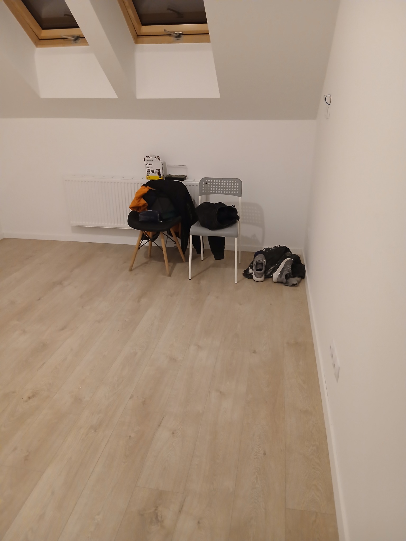 Jasne panele podłogowe w pokoju na poddaszu z oknami dachowymi. Na ścianie widoczny grzejnik i dwa krzesła z ubraniami. Neutralne kolory ścian i podłogi.