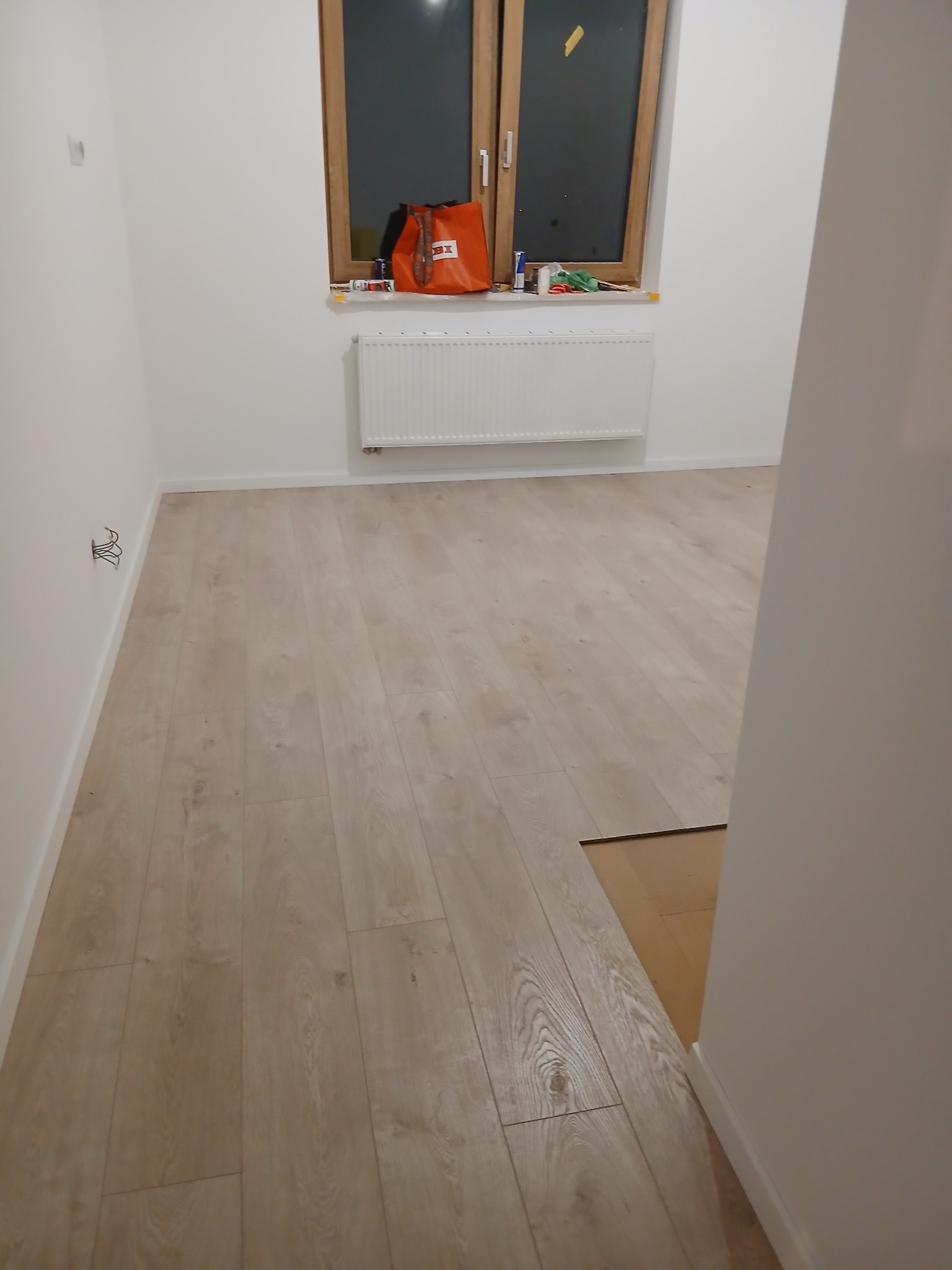 Jasne panele podłogowe ułożone w pokoju z białymi ścianami i oknem. Widoczny fragment nieukończonej podłogi przy ścianie. Torba i narzędzia na parapecie.