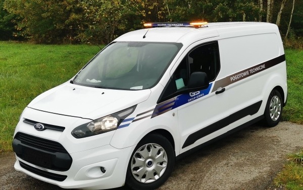 Biały furgon Ford Transit z napisem 'Pogotowie Techniczne' i lampą ostrzegawczą na dachu, zaparkowany na tle zieleni. Widoczne logo firmy GS Group-Serwis.