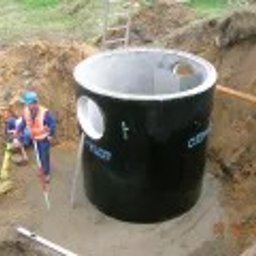 Futura Pro Iryna Pikula - Montaż betonowego zbiornika z otworem rewizyjnym w wykopie, widoczni pracownicy w odblaskowych kamizelkach i zagęszczarka wibracyjna. Etap instalacji.