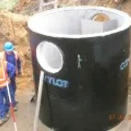 Futura Pro Iryna Pikula - Czarny, betonowy zbiornik Cylot z otworem, częściowo zagłębiony w ziemi. Dwóch pracowników w kaskach i kamizelkach odblaskowych w tle. Data w prawym dolnym rogu.