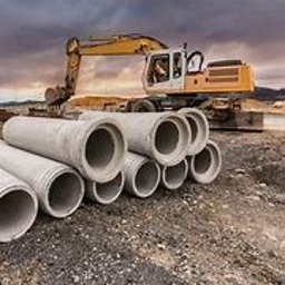 Betonowe rury kanalizacyjne ułożone na ziemi, z koparką w tle pod pochmurnym niebem. Widoczne żebrowania na rurach i nierówny teren.