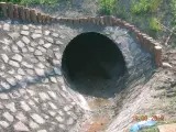 Betonowy przepust rurowy, wykończony kamienną obudową i ceglanym obrzeżem, odprowadzający wodę w terenie. Widoczne ślady wilgoci.