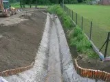 Wybetonowany rów melioracyjny w ziemi, zabezpieczony drewnianymi palisadami, z widocznym fragmentem ogrodzenia i zieleni w tle. Prace ziemne w toku.