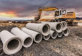 Betonowe rury kanalizacyjne ułożone na placu budowy, z koparką w tle i zachodzącym słońcem. Widoczne złączenia kielichowe rur.