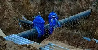 Zabezpieczone rury wodociągowe z niebieskimi zaworami w wykopie, widoczne rury na dnie wykopu. Etap budowy przyłącza wodociągowego.