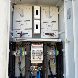 FABITECH - Otwarta skrzynka elektryczna z zamontowanym licznikiem i zabezpieczeniami. Widoczne przewody, listwy zaciskowe i elementy montażowe. Instalacja w trakcie podłączania.