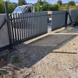 Nowoczesna, przesuwna brama wjazdowa z pionowych profili stalowych, częściowo otwarta, widoczny samochód dostawczy w tle, teren budowy.