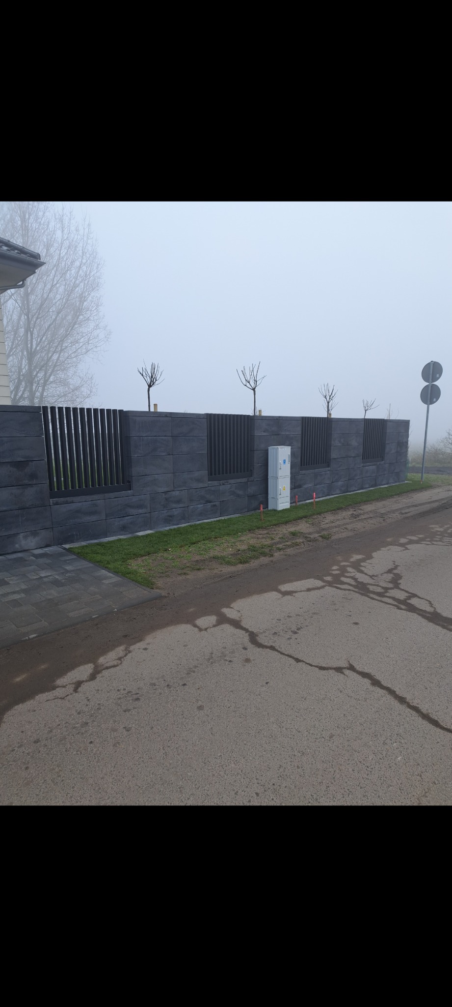 Nowoczesne ogrodzenie z szarego betonu architektonicznego z metalowymi przęsłami w pionowym układzie, widoczne na tle mglistego krajobrazu i skrzynki elektrycznej.