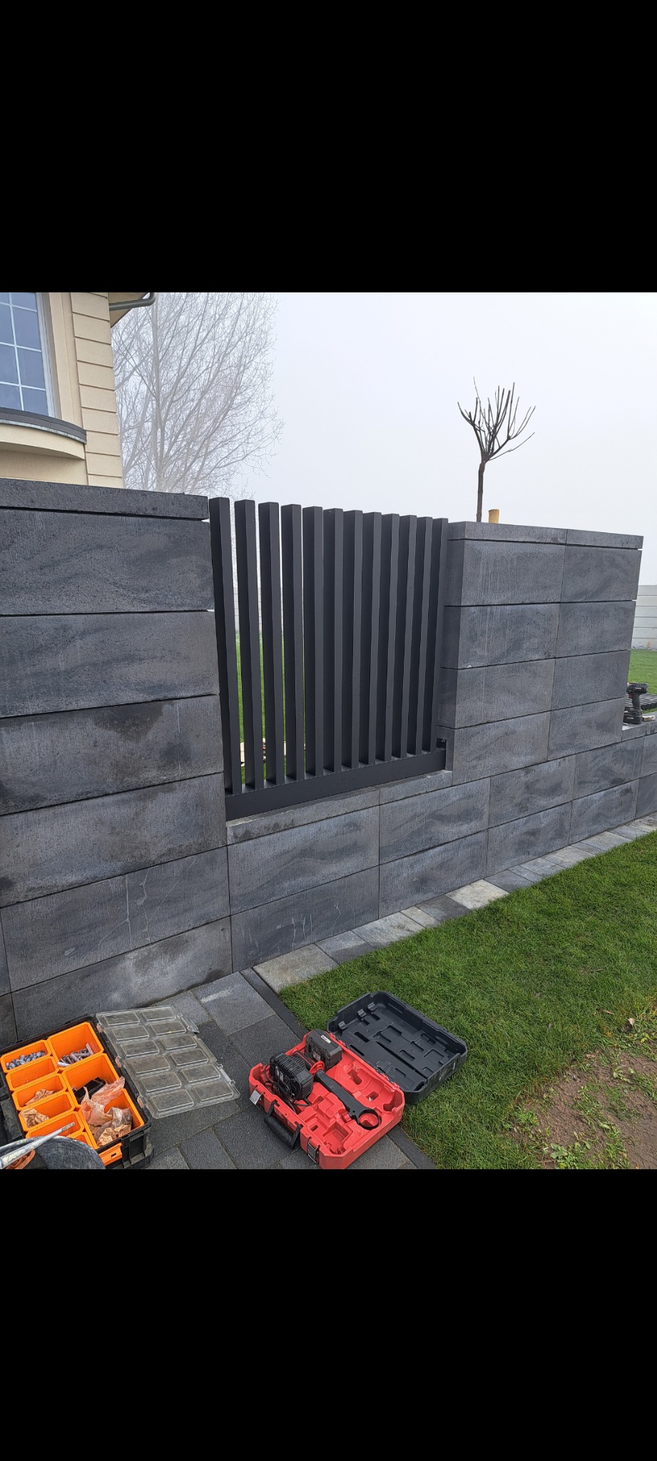 Montaż nowoczesnego ogrodzenia z betonowych bloczków i pionowych metalowych prętów, widoczne narzędzia i skrzynki z elementami montażowymi na kostce brukowej, trawnik w tle.