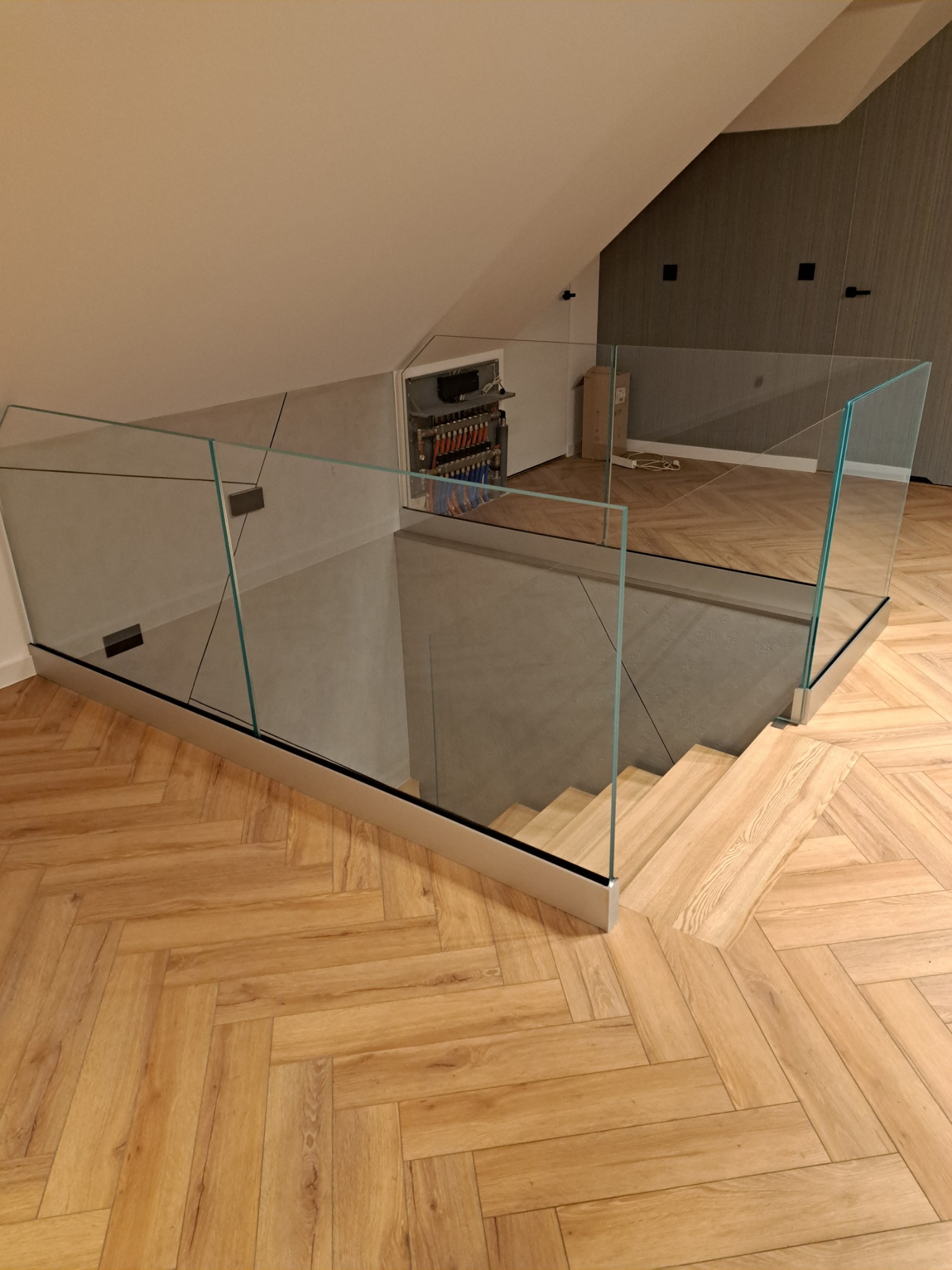 Szklana balustrada z metalowymi mocowaniami zabezpieczająca schody w nowoczesnym wnętrzu z parkietem ułożonym w jodełkę. W tle ściana z ciemną okładziną.